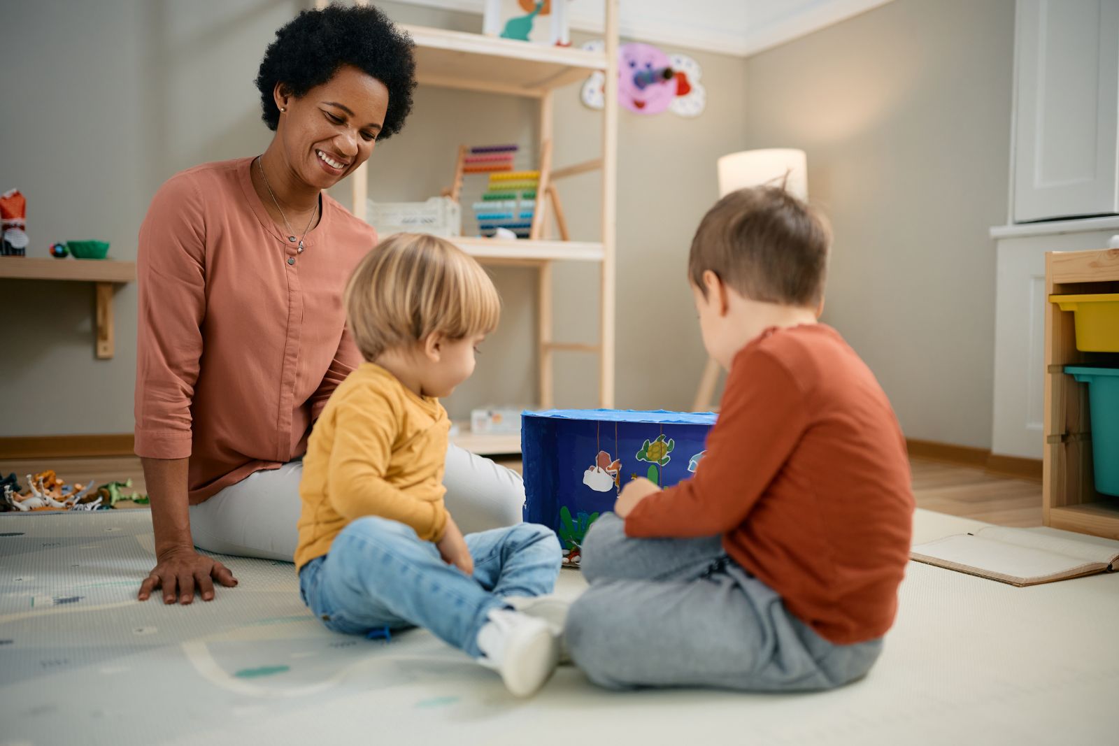 A carer smiling as two toddlers play with toys on the floor